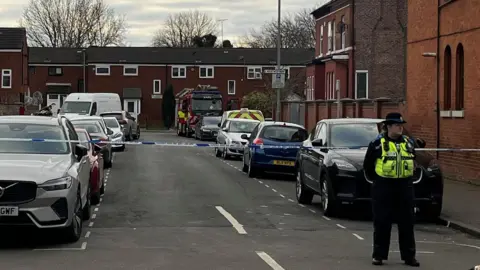 BBC A police officer stands in front of blue-and-white tape hanging across the road. A fire engine can be seen parked at the far end of Gateshead Close.