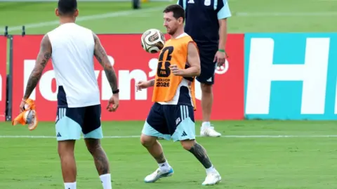 PA Media The image shows a football training session on a grass pitch. One player, Lionel Messi, wearing an orange training bib is controlling a ball in mid-air, while two other players in training kit stand nearby.