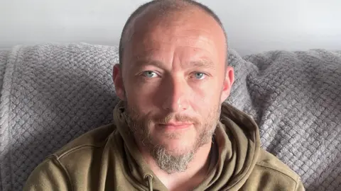 Robert has grey/blonde beard and a mostly shaven head. He is looking into the camera, wearing a brown/green hoodie. He is sitting on a sofa with a beige/grey thrown visible in the background.