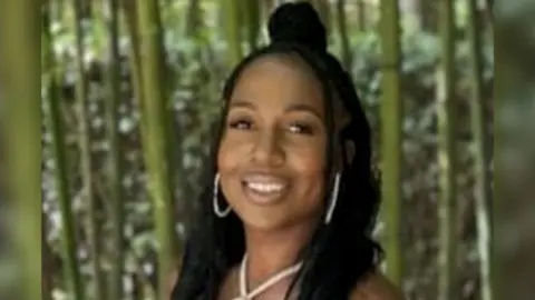 A young woman wearing gold jewellery smiles. She is standing in front of a forest. 