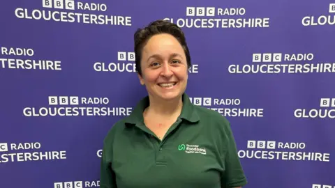 A smiling Vicky Ranford with her short dark brown hair wears a green polo shirt with the Gloucester Foodbank logo on the front. She is standing in from of the purple BBC Radio Gloucestershire branded back-drop.
