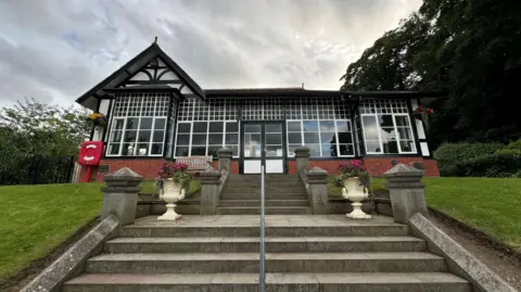 A black and white half-timber building at the top of two flights of steps. It is set within a park, with a lawned embankment in front and trees behind.