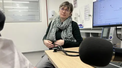 A patient talks to GP, with a foam-headed microphone recording their consultation. They are sat in a clinical room. The doctor is a woman with grey trousers and a stripy black and white scarf. She has short hair and large red-framed glasses. Only the right arm of the patient is visible