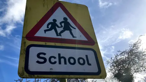 Nadia Lincoln/LDRS A rectangular yellow road sign against a blue sky. It depicts a red-outlined triangle showing a black silhouette of a girl leading a boy in profile on a white background. Beneath it is black bordered rectangle with School written on it in black letters. 