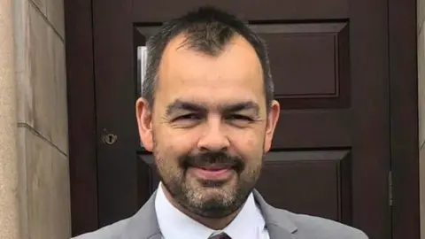 Stephen Kane Mr Kane with short black hair and facial hair. He smiles at the camera as he wears a grey suit with a white shirt and red patterned tie.