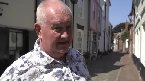LDRS A man with short grey hair and wearing a blue and white patterned shirt is standing in a narrow street with multi-coloured properties in the background.