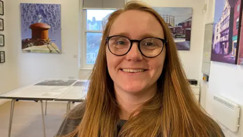 Steph Towns is a young woman with long auburn hair and glasses. She is standing in a room with pictures on the walls behind her and a window in the distance 