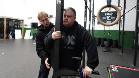 Patrycja Boryka/BBC A man is holding onto a black pole with his other hand on a black stand further down. He has short grey hair and a black hoody. A man next to him is assisting him by supporting him at his side and looking at him. Gym equipment can be seen in the background.