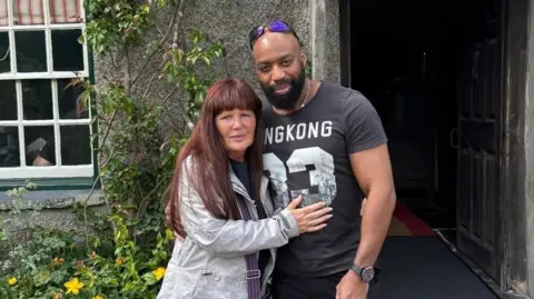 GMP Denise, with red hair and a light blue jacket, with her arms around her partner Sylvester Abayomi, wearing a black T-Shirt with white numbers and with sunglasses on his head, in a photo released by the family.