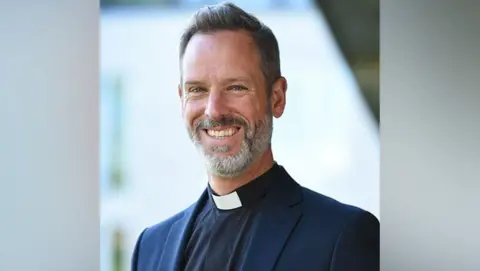 Ridley Hall Rt Revd Dr Michael Volland is facing the camera and smiling. He has a grey beard and dark hair and is wearing a dog collar and blue jacket.