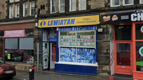 Google A row of shops on a town street, including a Polish food store with a bright yellow‑and‑blue sign.