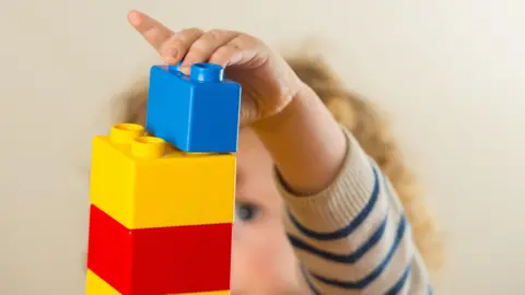 A child indoors playing with building blocks. The child's face is hidden but they have curly ringlet style blonde hair and are wearing a jumper which is beige with navy stripes up the arm. The child is building a tower out of yellow and red blocks and is pictured stacking a single blue block on the top of the tower.
