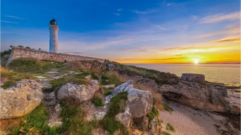 An orange sunset over the sea. There are rocks in the foreground and a lighthouse standing on the cliff.