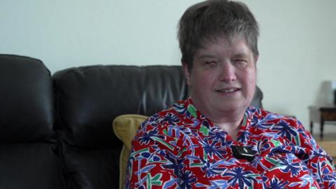An older brown haired lady wearing a flowery bright shirt and sat on on brown sofa