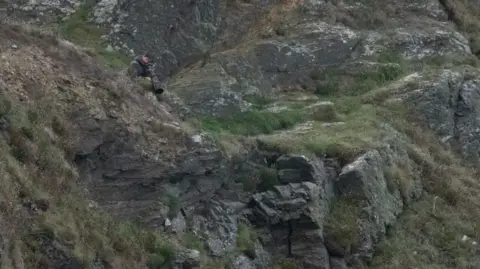 Martin Yelland A man using a telescopic camera lens on a clifftop to take a photograph of something below. He is wearing a camouflage jacket to blend in to the green and grey of the rock and grass behind him.