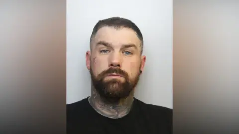 Avon and Somerset Police A mugshot of a man in his 30s with dark hair and a beard. He has tattoos all over his neck as well as one on one of his cheeks.