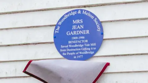 SIMON BALLARD A circular, blue plaque with white writing honouring Mrs Jean Gardner who is credited with saving Woodbridge Tide Mill from destruction. Gifting it to the town in 1977. The plaque is set against the white wall of the Tide Mill which is made up of horizontal planking. The cover that had been over the plaque hangs from the bottom of it.
