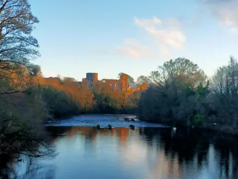 BBC Weather Watchers/Pop Pop A general view of Barnard Castle in the distance, taken from a point on the river looking down to the castle. The castle is hit by sunlight and its orange glow stands out from the rest of the area, which is in shadow. Trees line the river.