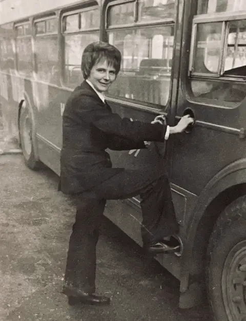BBC A black and white photo of Mary Curry standing next to a bus in 1973, one foot resting on the door. She is wearing a company uniform which includes a suit jacket and flared trousers. She has short hair.