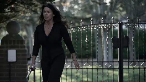 BBC/Left Bank Pictures/James Stack A woman with long dark hair wearing a black dress is walking in front of a iron gate holding a gun.