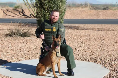 US Department of Homeland Security A man wearing tactical gear posing with a dog in a background that looks like desert terrain, with rocks and small shrubs