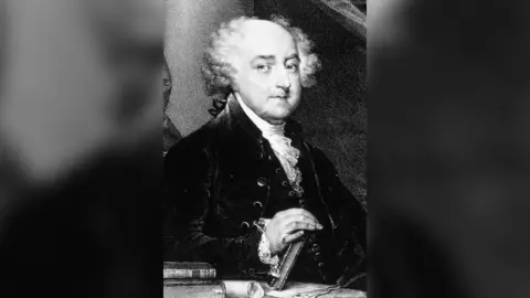 Getty/Hulton Archive A black-and-white portrait of John Adams. He is looking towards the right of the picture, wearing a dark jacket and white ruffled neckerchief. The top of his head is bald and he has white curly hair on the sides of his head. He is placing his hand on a book. 