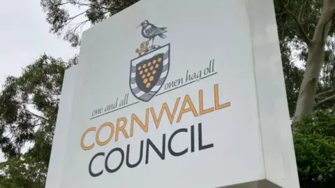 BBC Cornwall Council sign on a white background. There are trees in the background. The words are written in black and orange colours. 