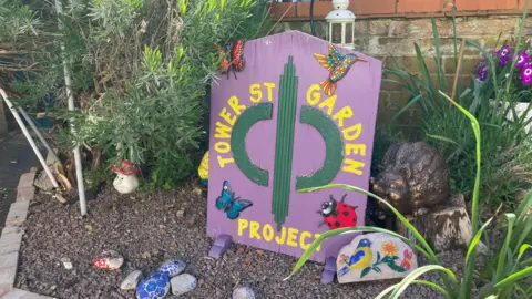 Shariqua Ahmed/BBC A purple Tower Street Project signboard with pebbles in front of it. It's placed within a front garden.