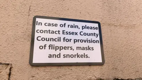 Lewis Adams/BBC A close-up picture of sign above an underpass that reads: "In case of rain, please contact Essex County Council for provision of flippers, masks and snorkels."