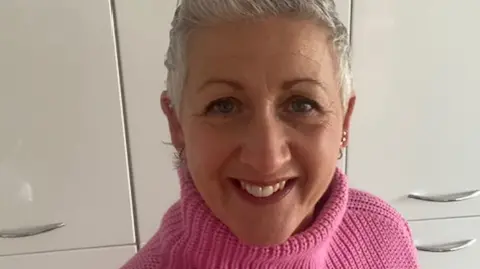 A close-up of Emma, wearing a pink polo-neck jumper, smiling at the camera with a fitted wardrobe behind her