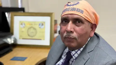 Steve Hubbard/BBC Jaswinder Kumar sitting down at a desk. He is wearing a grey suit jacket with a white and purple checked tie. He is looking at the camera. 