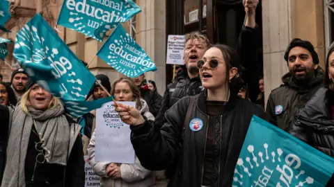 Getty Images Members of the NEU stand and wave green NEW flags as they protest demanding higher pay in London in February