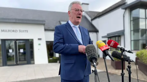 PA Media Mike Nesbitt wearing a blue suit and tie standing outside a building with white walls that has "Milford Suit" written above the entrance. He is standing speaking at some microphones.