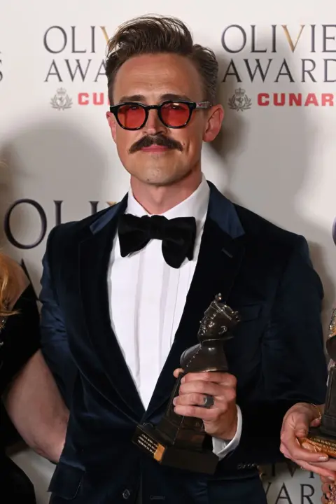 Getty Images Tom Fletcher in a suit and black bow tie wearing sunglasses and holding his Olivier award