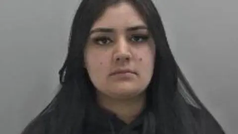 Sanaa Shahzad is a young woman who has long straight, dark hair and is seen wearing a black jumper. She has brown eyes and a mole on each cheek. 