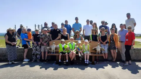 A large group of people, including four primary school children wearing high-visibility jackets, smiling around a wooden bench with a carving of an osprey on each end. It's a sunny day and the sky is blue.