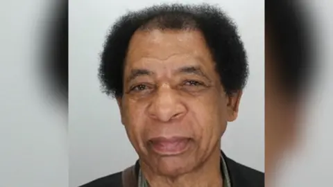 A mugshot of a man with short black hair looking into the camera in front of a grey background. 