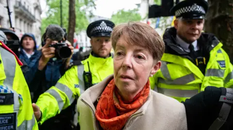 Getty Images Former Post Office chief executive Paula Vennells, wearing an orange scarf and tan jacket, flanked by police in hi-vis jackets.