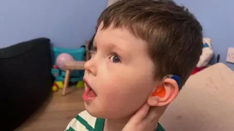 A young boy with short brown hair turns his head to show his orange and blue hearing aid in his ear. He is wearing a green and white t-shirt and is standing in a blue painted room with toys on the floor.