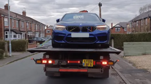 A blue sports car is hauled away on a truck through a residential area. 