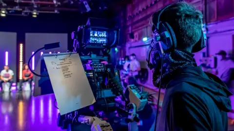 National Film and Television School In a TV studio, a camera operator with a headset works beside a camera that has a cue sheet labelled ‘Camera 1’
