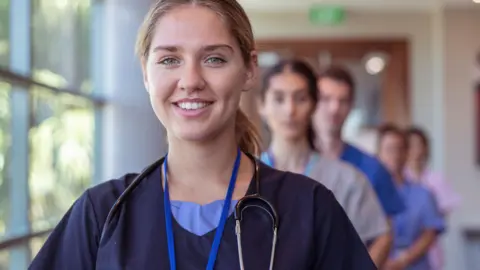 Health professionals standing in a line in a hospital corridor, with a woman at the front smiling at the camera.