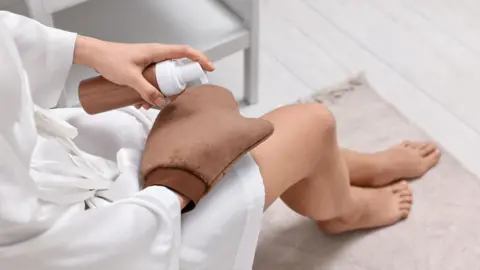 Getty Images A generic image of a woman wearing a brown mitt and holding a fake tan bottle. Her face can't be seen. She is wearing a white dressing gown and has her legs and feet on the rug.