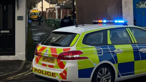 A photo of a police car parked in front of an alleyway. There is an ambulance in the background and a number of emergency workers