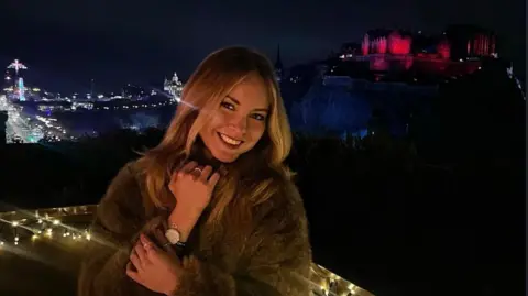 Mira Al Faradz A photo with Anastasiia Korkina on the foreground and Edinburgh castle on the background. The photo is made at night and the Edinburgh castle is lit with pink light.