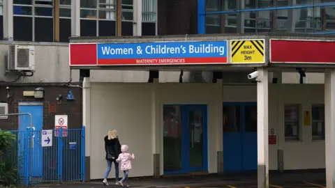 PA Media An unidentified blonde-haired woman wearing a black coat walks hand-in-hand with a young girl in a pink coat. They both have their backs to the camera as they make their way towards the entrance of the Women & Children's Building at the Countess of Chester Hospital, which is marked with a large blue sign.