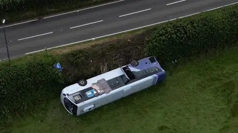 A bus overturned on grass near a main road 