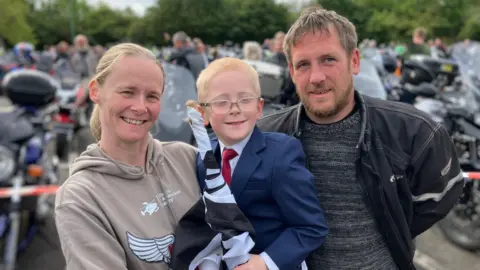 BBC A smiling Teddy, who is wearing glasses and a dark blue suit jacket, and red tie. He is being held by his mother who is wearing a beige hoodie with blond hair, tied back. His father on the other side has a beard and is wearing a biker jacket, jumper and orange trousers. 
