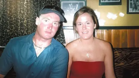 Lucy Early A man in a blue shirt and wearing a cap on the left is next to a woman wearing red on the right. They are both looking at the camera and near curtains and a yellow and brown wall.
