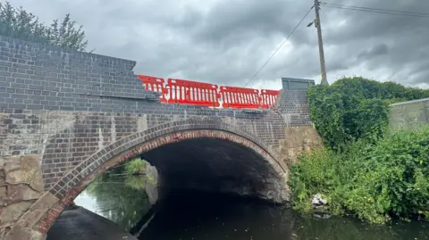 A bridge over a canal with many bricks missing due to a vehicle strike. Orange safety barriers can be seen on top of the bridge.